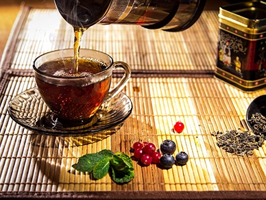 A cup is being filled with tea. Berries and leaves are seen on bamboo table mat.